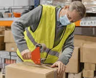 Trabalhador aplicando fita adesiva industrial para fechar uma caixa em um ambiente de armazenamento.
