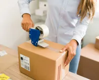 Pessoa fechando uma caixa de papelão com aplicador de fita adesiva transparente em um ambiente de trabalho.