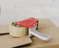 Aplicador manual com fita adesiva industrial sobre caixa de papelão, utilizado em processo de embalagem logística.