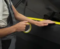 Aplicação de fita adesiva para proteção de superfície em veículo, utilizada para evitar riscos durante processo industrial.