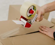 Fita adesiva industrial sendo aplicada em caixa de papelão para fechamento e vedação de embalagens no ambiente logístico.