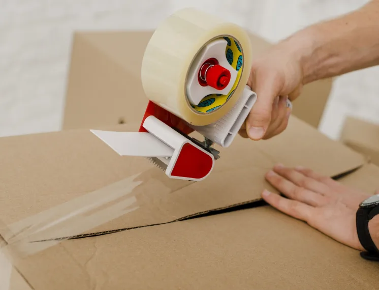 Fita adesiva industrial sendo aplicada em caixa de papelão para fechamento e vedação de embalagens no ambiente logístico.