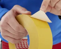 Mãos aplicando fita adesiva industrial dupla face amarela em um rolo, com o liner sendo removido.